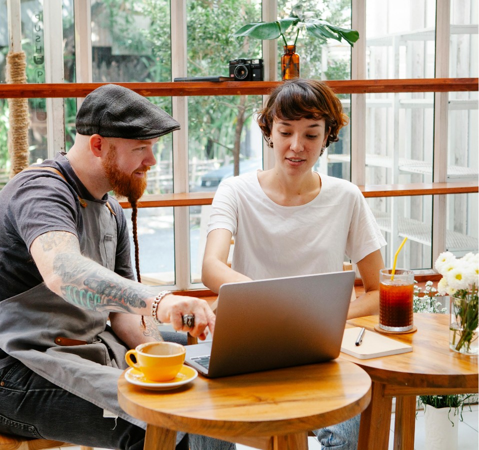 coffee shop with a man in an apron sitting with a woman in front of a laptop discussing